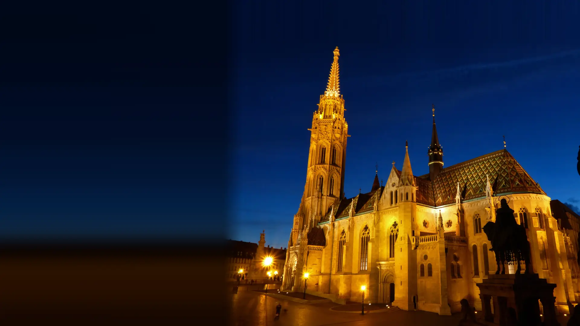 Matthias Church in Budapest at night, floodlit in gold under a deep blue sky, with its tall spire and its roof of coloured diamond-pattern tiles